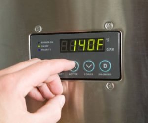 "A male finger is turning up the temperature on a tankless water heater control panel. Tankless water heaters are new technology which produces hot water on demand and is much more energy efficient than a conventional hot water holding tank system. This unit is installed and in operation, there are some scratches, dust and scuff marks on the surface."